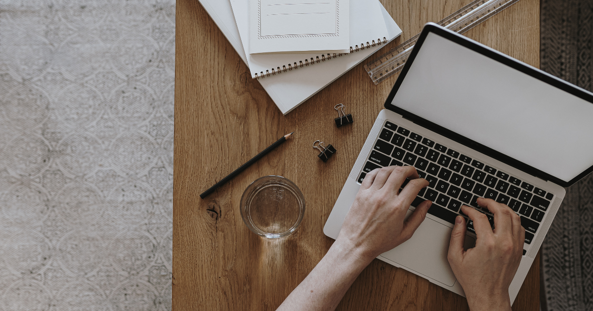 On a desk, there is a laptop, binder clips, notepads, a pencil, a ruler, and a glass of water.  A person is typing on the laptop.